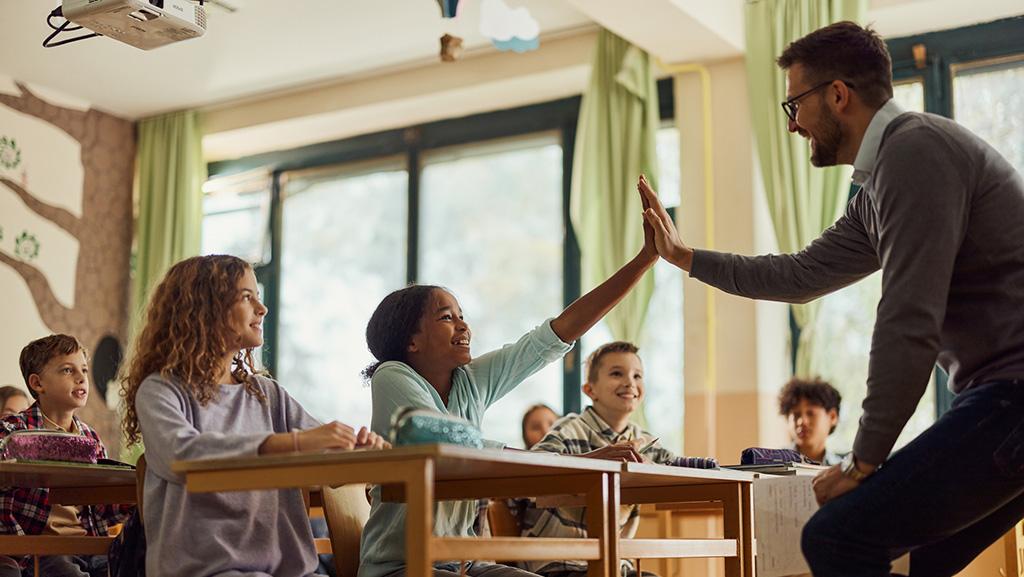 Rigor and relevance teacher high fiving student in classroom and the rest of the sudents are engaging in instruction